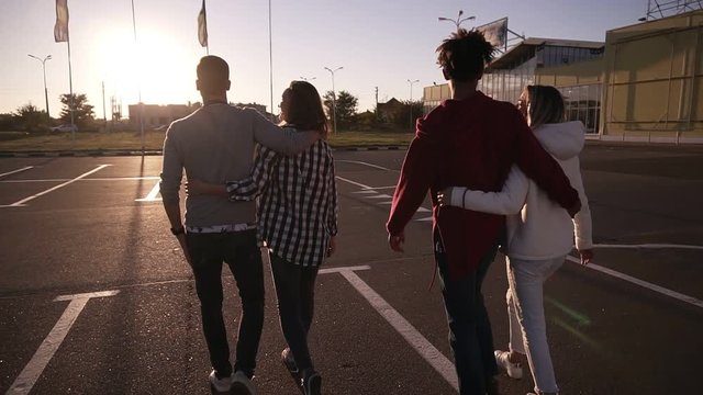 Group of young multirace couples laughing while walking by asphalt road. Young men and women enjoying a summer day at parking zone near the mall. Front view, Sun shines on the background. Rare view