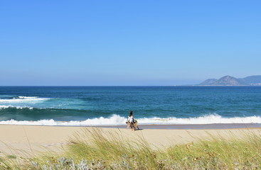 Fototapeta na wymiar Wild beach with women and dog walking. Bright sand, vegetation in sand dunes and blue water with waves and foam. Blue sky, sunny day. Galicia, Spain.