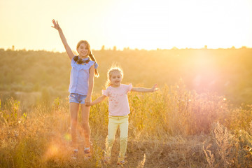 Fototapeta premium Little smiling girls stand on sunshine evening field with joyfully raised hands