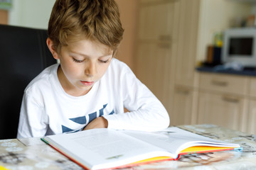 Little blonde school kid boy reading a book at home. Child interested in reading magazine for kids. Leisure for kids, building skills and education concept..