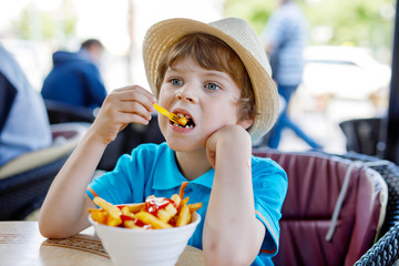 Cute healthy preschool kid boy eats french fries potatoes with ketchup sitting in cafe outdoors....