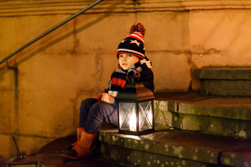 Fototapeta na wymiar Little cute kid boy with with a light lantern on stairs near church. Happy child on Christmas market in Germany. Kid waiting on parents on cold winter day