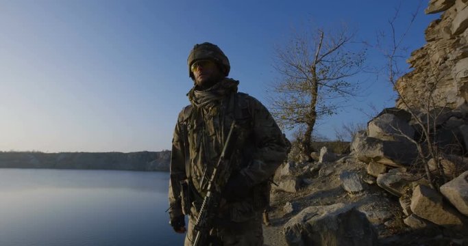 Medium slow motion shot of equipped and armed soldiers walking in single file by the side of a lake