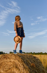 Naklejka na ściany i meble Cheerful young girl standing on a haystack in the middle of a wheat field looking into the distance
