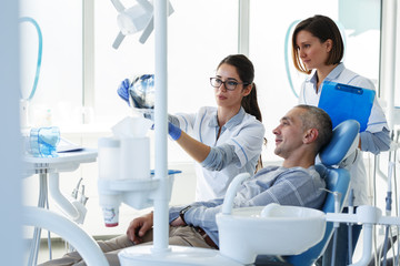 Dentist and her assistant in dental office talking with male patient and preparing for...