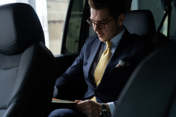 Young businessman sitting on back seat of the car, while his chauffeur is driving automobile. - obrazy, fototapety, plakaty