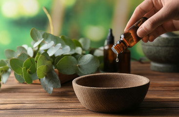 Woman pouring eucalyptus essential oil into bowl on wooden table - obrazy, fototapety, plakaty