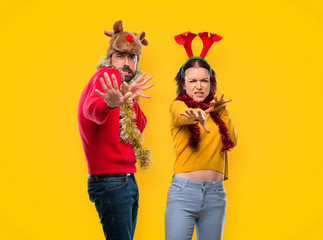 Couple dressed up for the christmas holidays is a little bit nervous and scared stretching hands to the front on yellow background