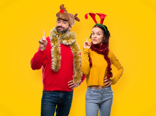 Couple dressed up for the christmas holidays showing and lifting a finger in sign of the best on yellow background