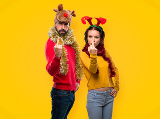 Couple dressed up for the christmas holidays making horn gesture. Negative expression on yellow background