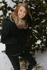 Cute Young Blonde Caucasian Woman with Scarf Outside Enjoying the Fresh Winter Snow on the Pine Trees