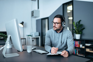 Tech manager with headset writing on the clipboard  and looking at computer screen.