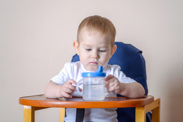 Baby boy drinking water studying a plastic bottle while sitting on a chair in the kitchen.