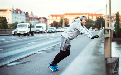 Mature male runner with headphones stretching outdoors on the bridge in Prague city.
