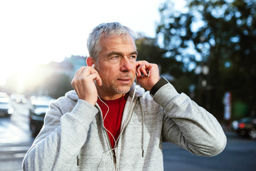 A portrait of an active mature man with earphones standing outdoors in city.