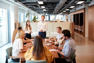 Businessman And Businesswoman Addressing Group Of Young Candidates Sitting Around Table And...
