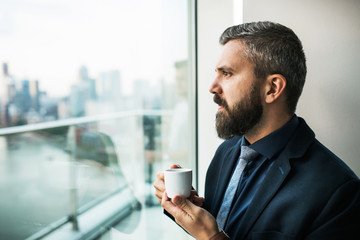 A portrait of businessman with a cup of coffee looking out of a window in an office.