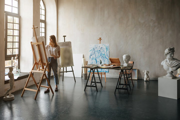 Artist at the working process. Young woman creating the painting. Workshop room with light and...