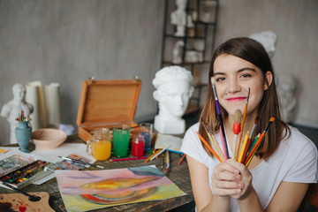 Adorable young artist girl holding brushes and smiling. Creative workshop room at the background
