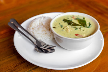 Green curry with red chili chicken and green basil leaf in the white bowl with steamed rice in the white round plate with fork and spoon on the brown wooden table. 