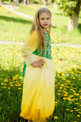 Blond young girl posing in a yellow green dress standing on the grass with dandelions yellow flowers