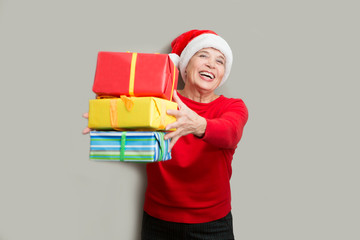 Happy senior lady in Santa red hat with Christmas presents looking at camera and smiling posing about a wall.  Christmas time.