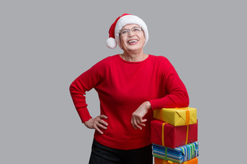 Happy senior lady in Santa red hat with Christmas presents looking at camera and smiling posing about a wall.  Christmas time.