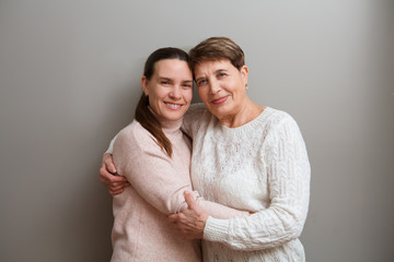 Happy senior lady mother and adult daughter embracing looking at camera on a gray background