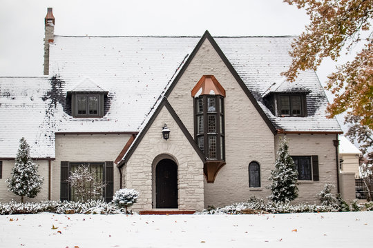 Charming white painted brick cottage with copper accents during snowfall with autumn leaves still on trees