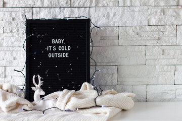 Baby it's cold outside phrase made with white letters on a black wooden board with lights and with a deer statuette on white background