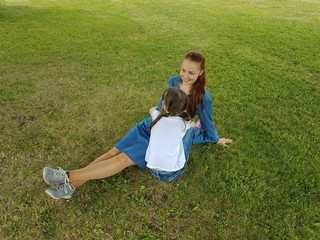 a beautiful young woman in a blue denim dress is sitting with her daughter in the Park under a big tree on a summer day.