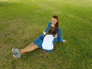 a beautiful young woman in a blue denim dress is sitting with her daughter in the Park under a big tree on a summer day.