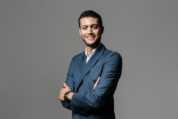 Image of optimistic arabic businessman 30s in formal suit looking at camera standing with arms crossed, isolated over gray background