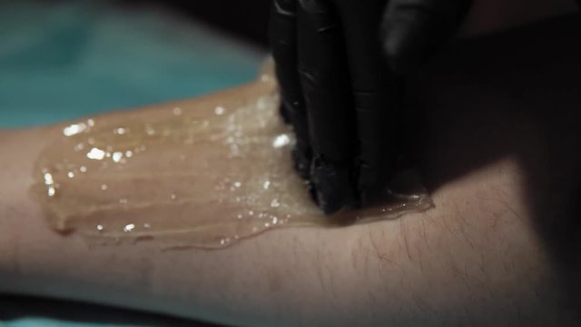 A sugaring is performed using the sugar paste from the legs of a young woman at a spa. The master in black gloves removes the hair in the direction of growth.