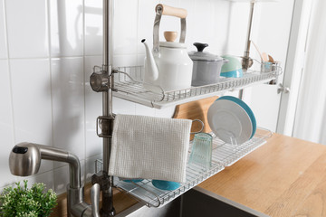 Clean dishes drying on metal dish rack on light background. Kitchen utensils and dishware on wooden shelf. Kitchen interior background.Text space.