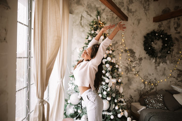 Charming girl dressed in white sweater and pants stands next to the New Year tree in front of the...