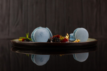 Blue macarons with blackberries and raspberries on beautiful plate, on the mirror table, reflection, black table, black background