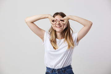 Fototapeta premium Funny girl dressed in a white t-shirt and jeans makes glasses with her hands on her eyes on a white background in the studio