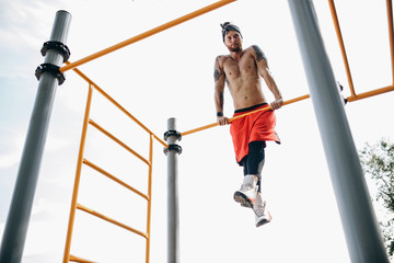 Athletic guy in headband with naked torso dressed in black leggings and red shorts makes the rise on the bar on the sports ground outside