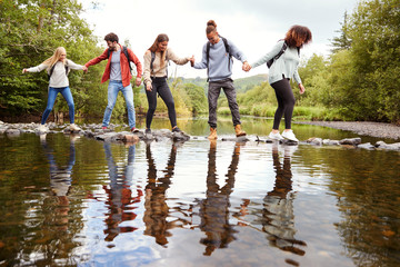 Multi ethnic group of five adult friends hold hands and help each other while carefully crossing a stream standing on stones during a hike