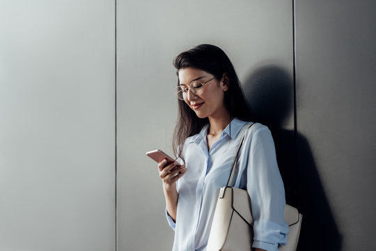 Pretty Thai smiling woman standing by the grey wall holding a purse and looking at her smart phone.