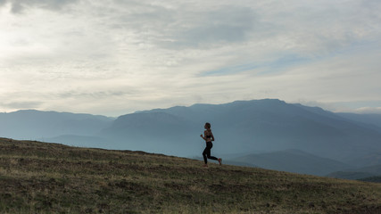 Fototapeta na wymiar Slim girl wearing black sport outfit is jogging in mountains, beautiful landscape, cloudy sky, foggy hills.
