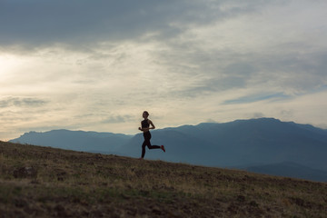 Slim girl wearing black sport outfit is jogging in mountains uphill, beautiful landscape, cloudy sky, foggy hills, sun is shining
