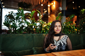 Pretty indian girl in black saree dress posed at restaurant with mobile phone at hand.