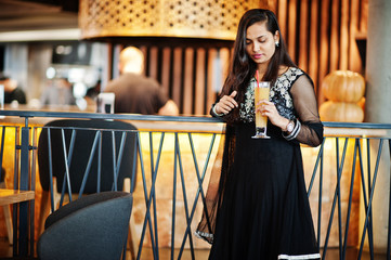 Pretty indian girl in black saree dress posed at restaurant with orange juice at hand.