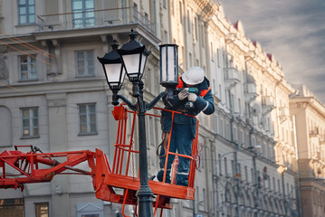 Worker at height in basket of hydraulic crane drills street lamp to replace cover. Professional...