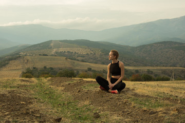 Naklejka na ściany i meble Smiling young girl is squatting crosslegged among dried yellow grass, meadow, in mountains, cloudy sky