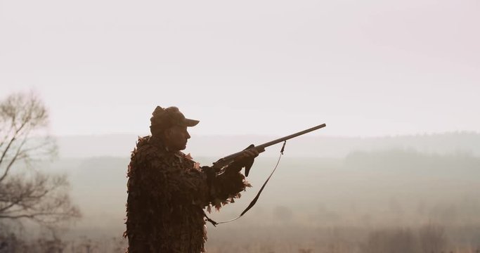Portrait of hunter in hunting equipment. Silhouette of man in sunset light aim with rifle foggy landscape on background