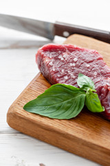 Raw meat with marinated salt, pepper and Basil seasoning leaf. Morning in the kitchen. Raw fresh beef steak on a wood cutting board. White wooden background, top view, copy space, Daylight