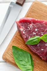 Raw meat with marinated salt, pepper and Basil seasoning leaf. Morning in the kitchen. Raw fresh beef steak on a wood cutting board. White wooden background, top view, copy space, Daylight
